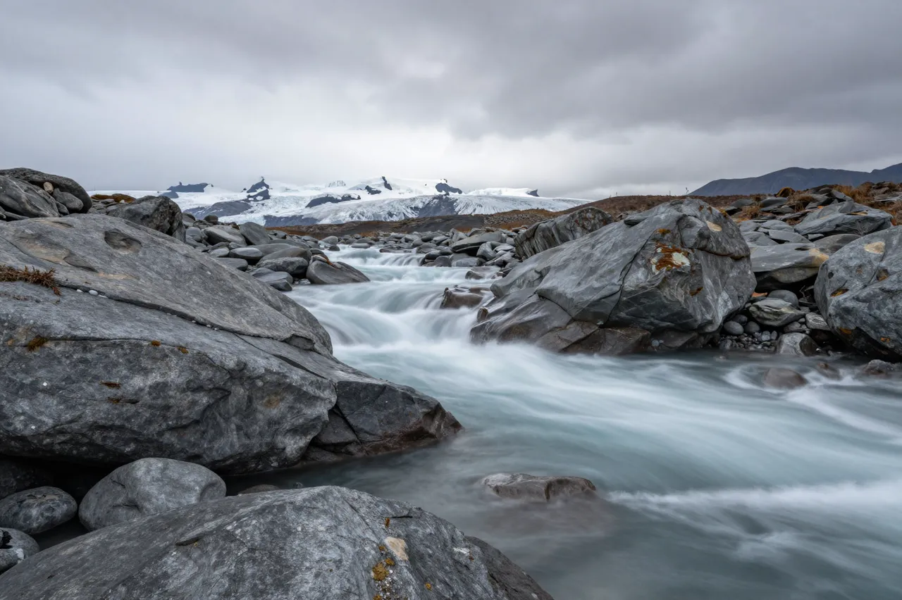 Long exposure shot of a milky glacial river flowing between gray boulders with distant glaciers under low heavy clouds