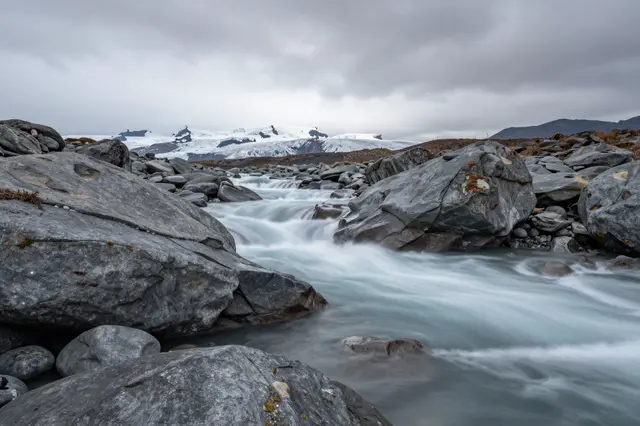 Long exposure shot of a milky glacial river flowing between gray boulders with distant glaciers under low heavy clouds