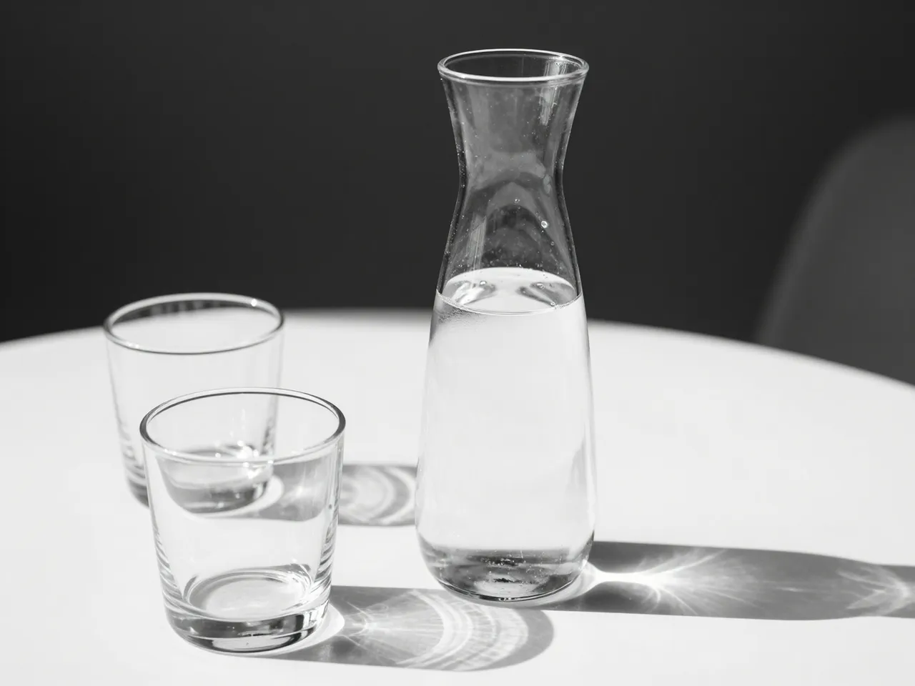 Black and white still life of a glass water carafe and two tumblers resting on a white table in sharp hard sunlight.