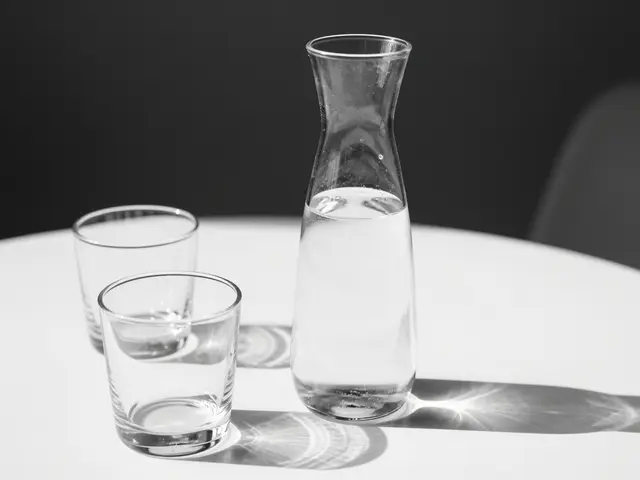 Black and white still life of a glass water carafe and two tumblers resting on a white table in sharp hard sunlight.