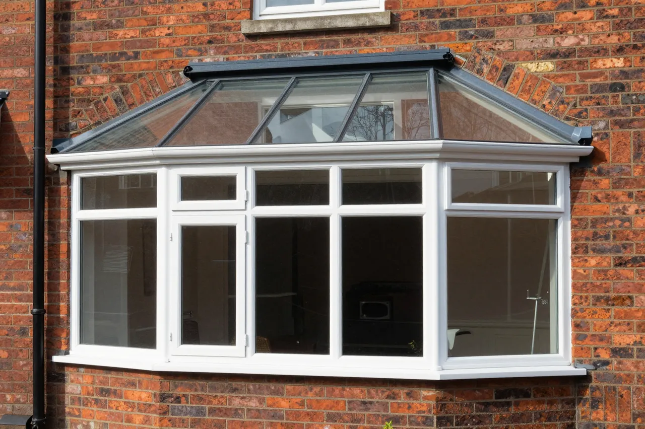 A white framed glass conservatory bay window attached to a red brick residential house in bright sunlight
