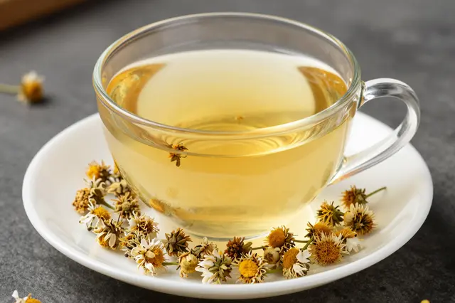 A clear glass cup of pale chamomile tea sits on a white saucer scattered with dried chamomile flowers.