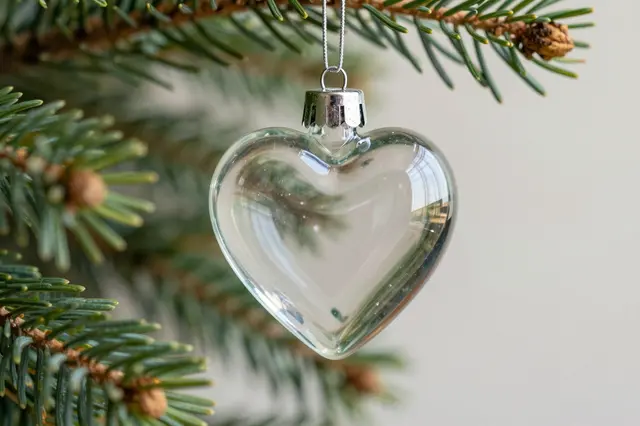 A clear glass heart ornament hangs from a pine tree branch against a soft neutral background