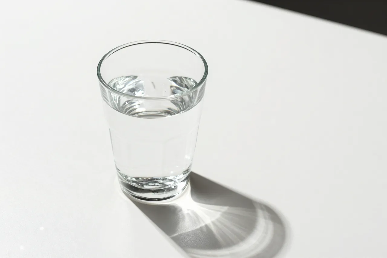 A clear glass of water sits on a white table casting bright caustic light patterns, with empty copy space to the right.