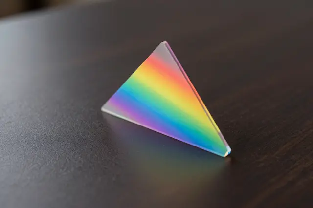 A triangular glass prism resting on a dark wooden table displays a rainbow light spectrum.