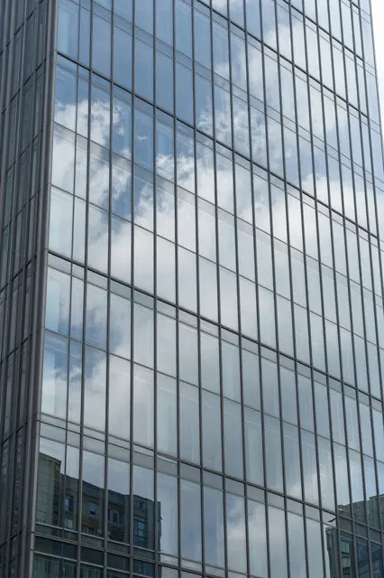 Angled close-up of a modern glass skyscraper facade reflecting fluffy clouds and surrounding city buildings