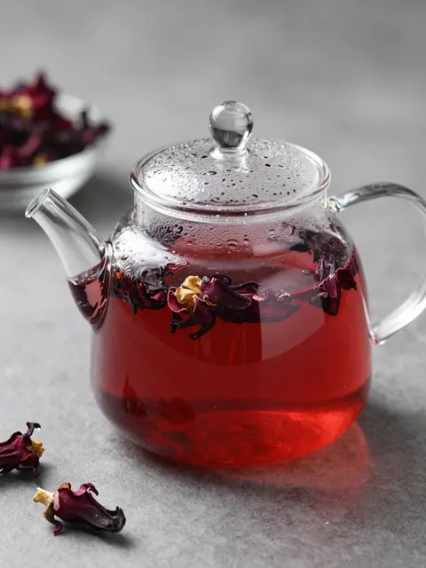 Clear glass teapot of deep red hibiscus tea with dried flowers rests on a gray matte surface
