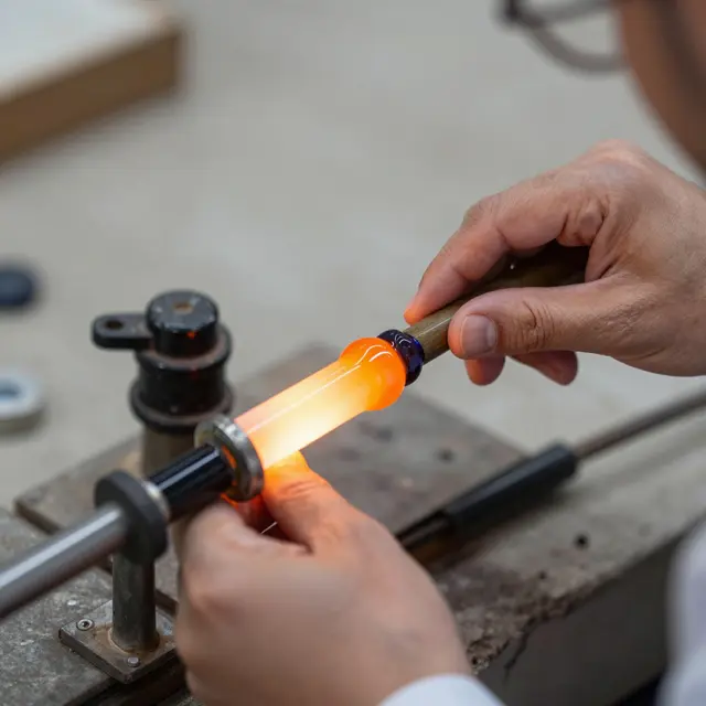 Close-up of a glassblower's hands working a pipe holding glowing orange molten glass at a workshop workbench