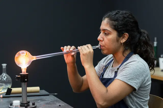 A woman blows through a glassblowing pipe to shape a glowing molten glass bubble in a dark workshop