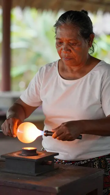 A mature Pacific Islander woman works on a glowing hot glass piece at an outdoor glassblowing station