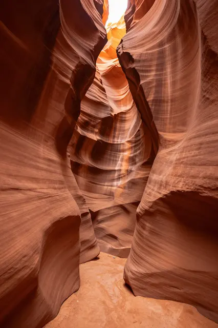 Smooth layered sandstone walls of a narrow Arizona slot canyon glow with warm orange light from above