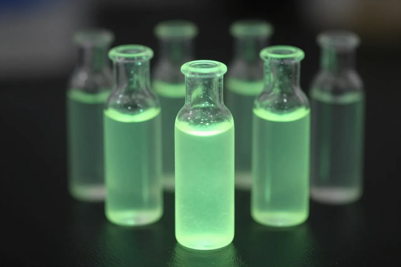 Glass vials holding bright glowing green chemiluminescent liquid sit arranged in a dark lab setting
