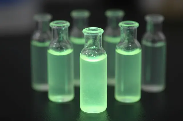Glass vials holding bright glowing green chemiluminescent liquid sit arranged in a dark lab setting