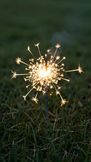 A lit golden sparkler stands upright in dark grass, emitting bright starburst sparks against a blurred lawn background.