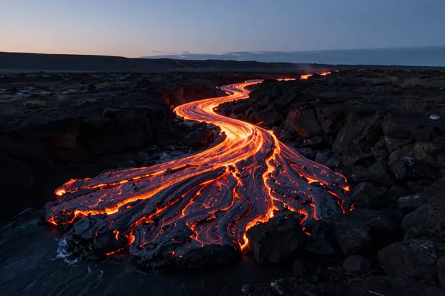 A winding glowing river of lava flows across dark volcanic rock toward water during twilight dusk