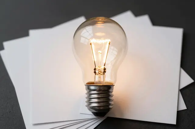 A lit vintage glass light bulb rests on fanned blank white paper cards against a dark background