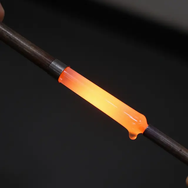 Bright glowing molten orange glass droops from the end of a glassblowing blowpipe against a dark background