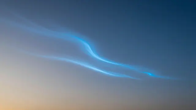 Wispy electric blue noctilucent clouds drift across a gradient twilight sky after sunset