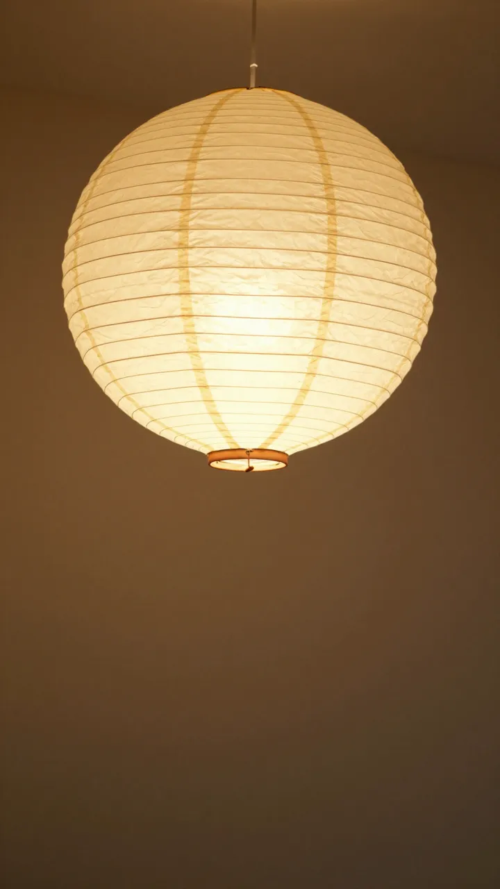 A round glowing white paper pendant lantern hangs against a dim warm brown wall with empty space below.