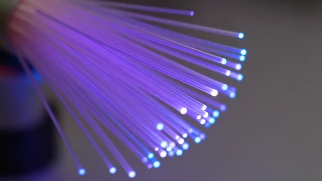 Close-up of fanned fiber optic strands emitting purple and blue glowing light against a soft gray background