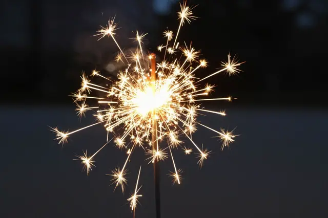 A lit sparkler bursts with bright golden sparks against a dark blurred evening background.