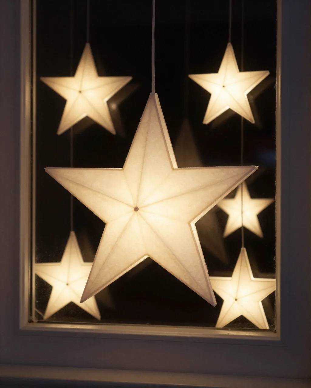 Warm lit white paper star lanterns hang inside a dark window for Christmas