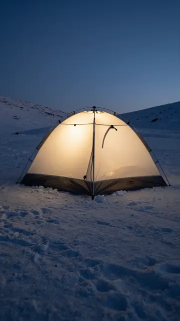 Warmly lit camping tent sits on a snowy mountain slope under a clear dusk sky