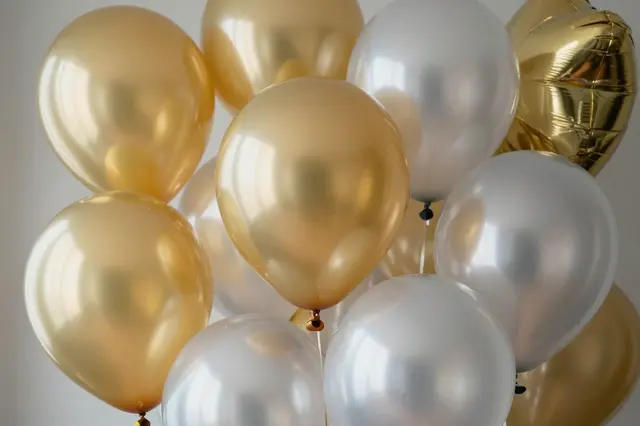A cluster of metallic gold and silver balloons arranged against a plain light neutral wall.