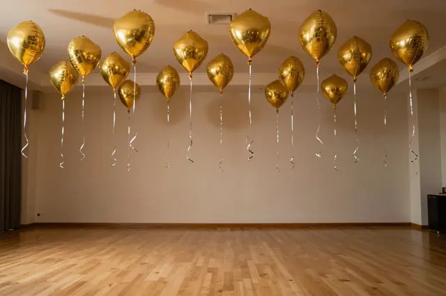 Shiny gold foil balloons hang with silver ribbons above an empty polished wood ballroom dance floor.