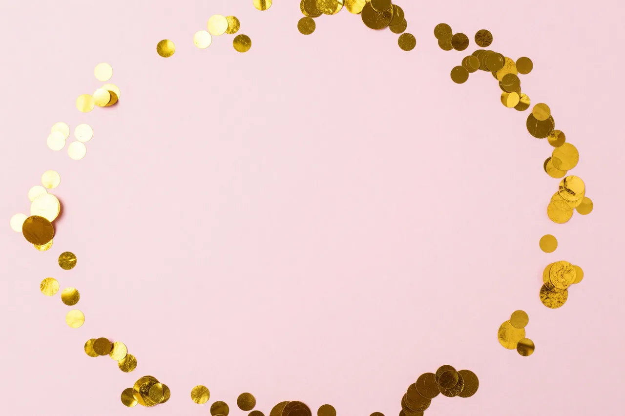 A top view of round gold confetti arranged in a loose circular frame on a pale pink background with empty central copy space.