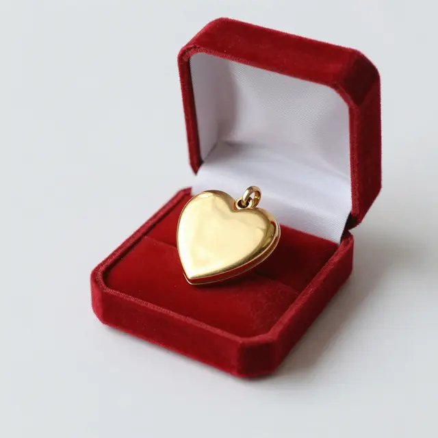 A polished gold heart locket rests inside an open red velvet jewelry box against a plain light gray background.
