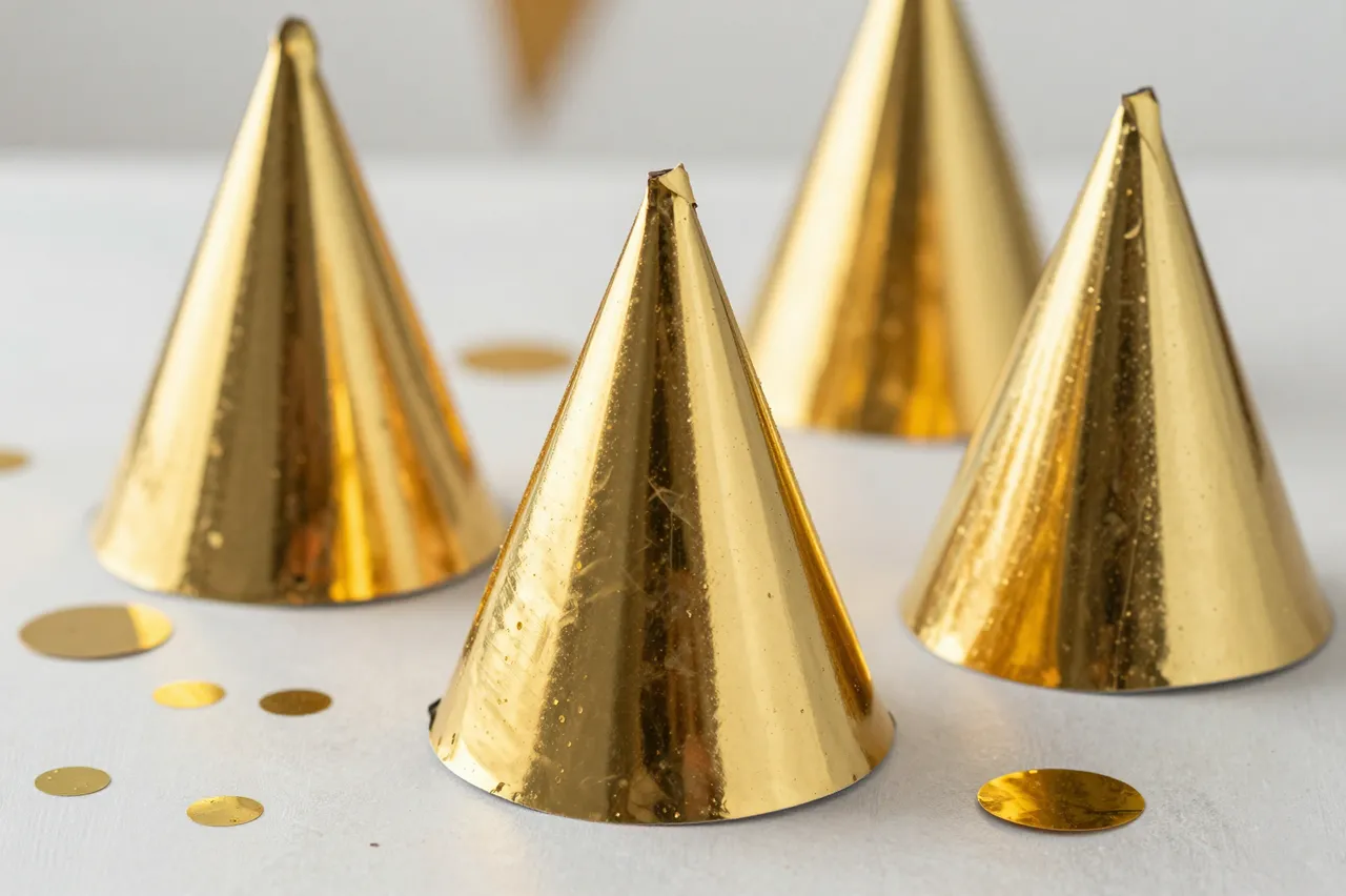 Shiny metallic gold party hats and scattered gold confetti rest on a pale tabletop.