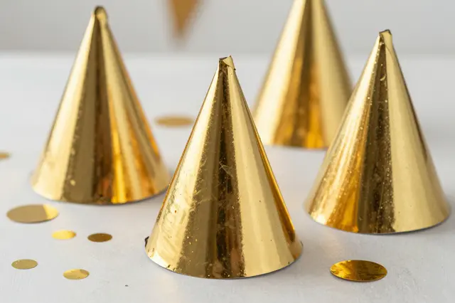 Shiny metallic gold party hats and scattered gold confetti rest on a pale tabletop.