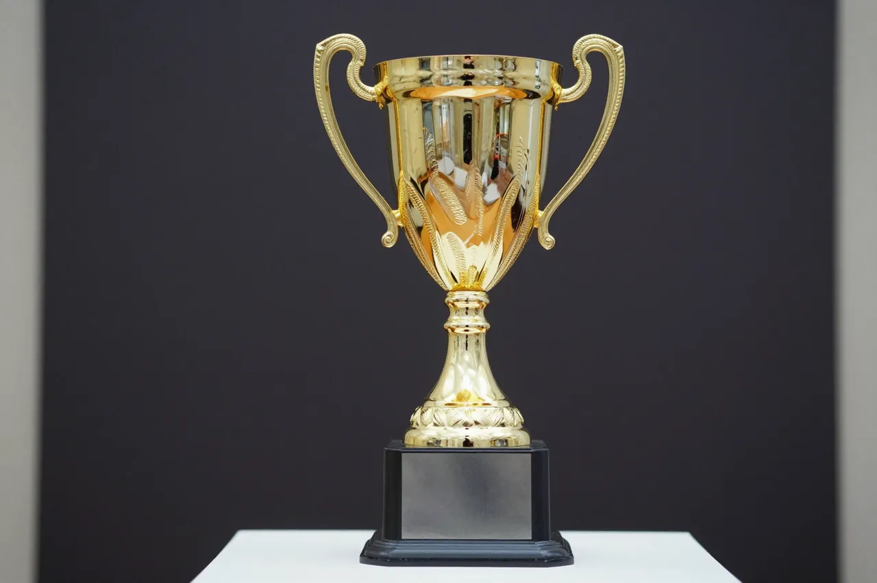 A shiny golden trophy cup rests on a dark base against a plain dark background