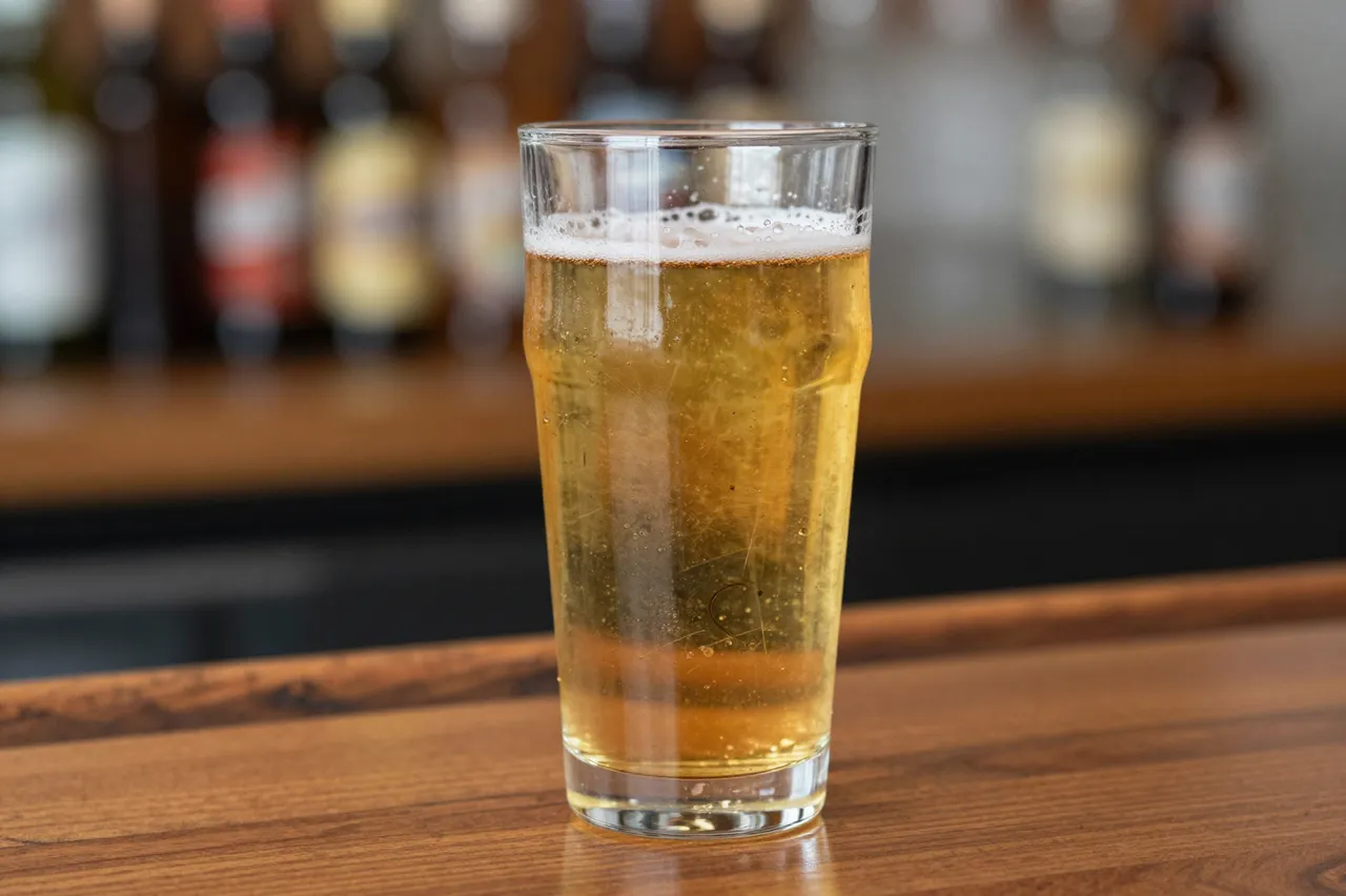 A full glass of frothy golden beer sits on a polished wooden bar top with blurred liquor bottles in the background.