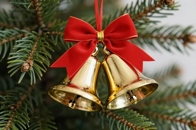 Shiny golden Christmas bells tied with a red bow hang from evergreen pine branches.