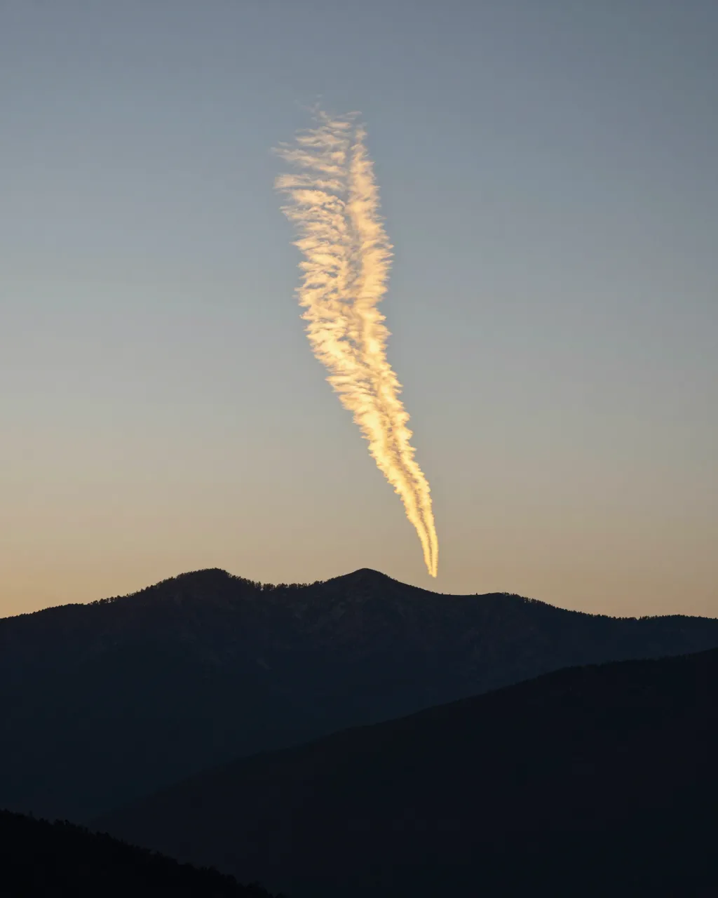 Glowing golden airplane contrail hangs above dark silhouetted mountain peaks at sunset