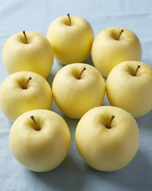 Eight pale yellow Golden Delicious apples rest on a soft pale blue linen cloth under gentle studio lighting