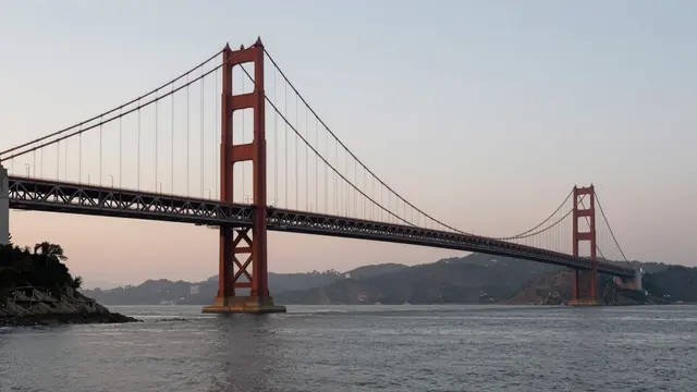Reddish suspension bridge spans a calm bay under a soft gradient dusk sky with distant hills