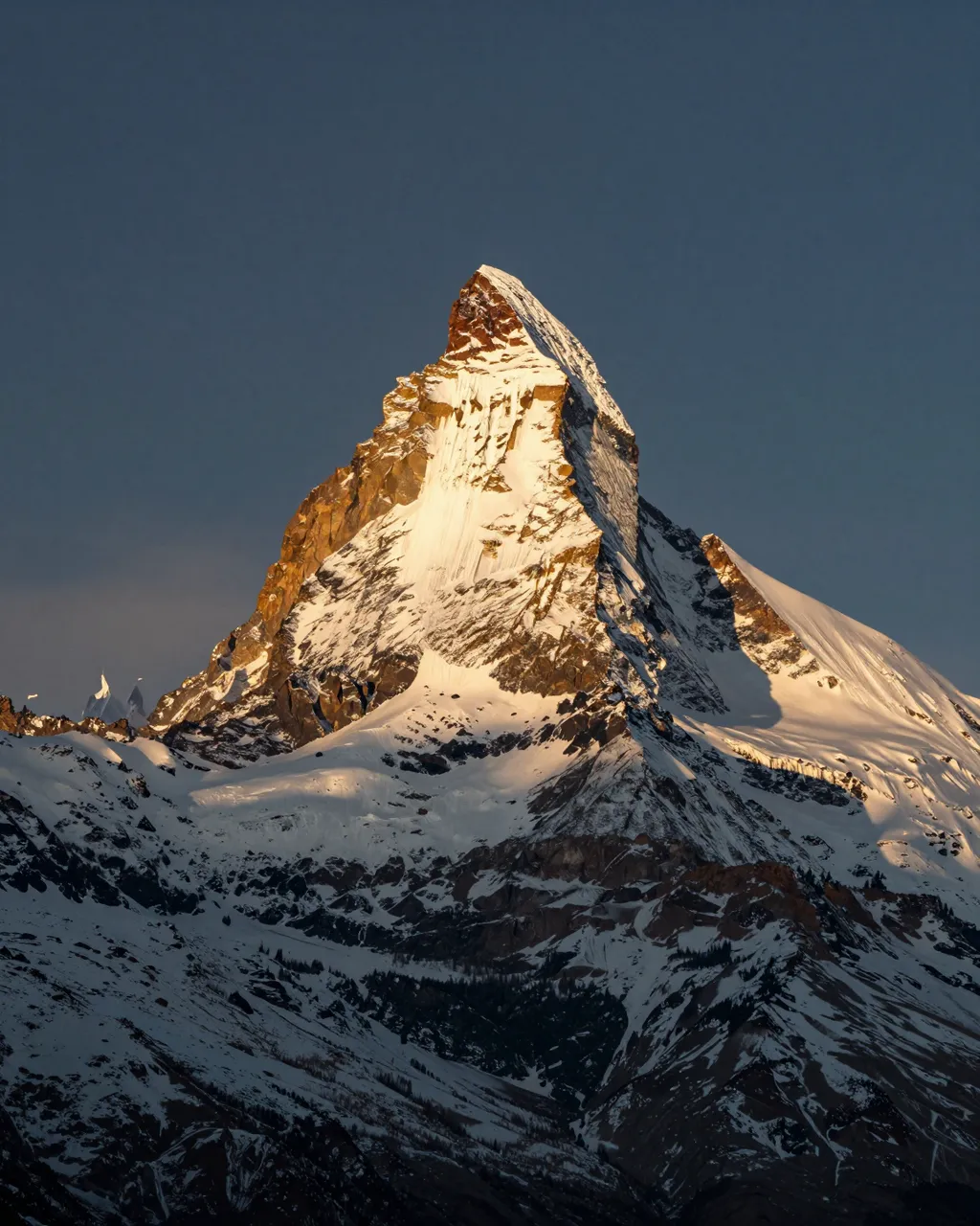 A sharp snowy alpine peak catches golden sunrise light above shadowed lower valley slopes under a dark clear sky.