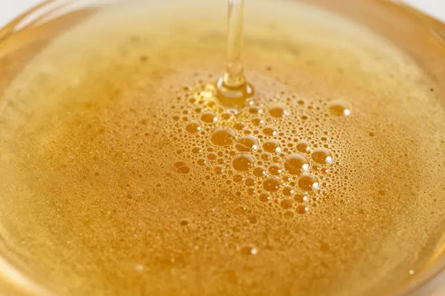 A thin stream of golden honey pours into a glass container, creating a surface dotted with varying sized bubbles