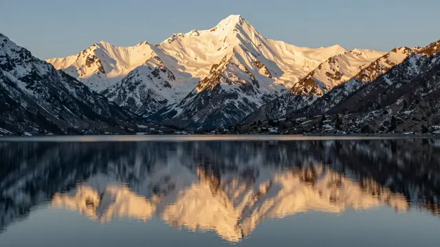 Sunlit snow-capped mountain range is reflected in a perfectly calm alpine lake at golden hour