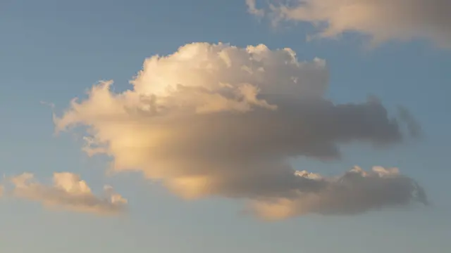 Fluffy cotton clouds glow with warm golden light against a soft pale blue sky at golden hour