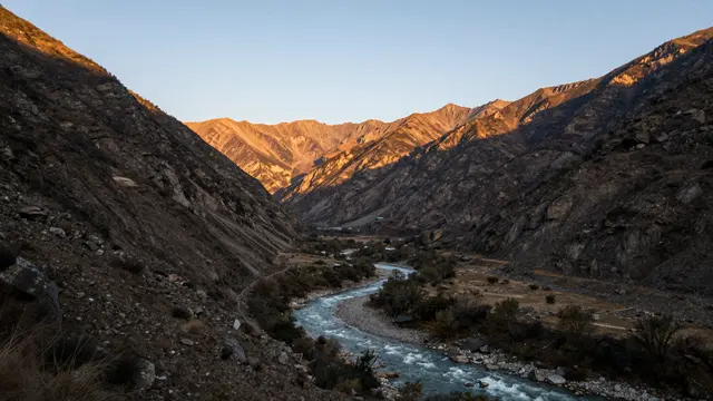 Sunlit distant mountain peaks overlook a winding river flowing through a rocky alpine valley at golden hour