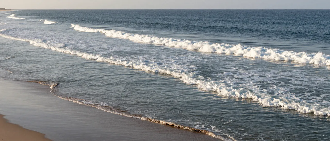Parallel lines of foamy ocean waves roll gently toward a quiet sandy beach in soft warm light