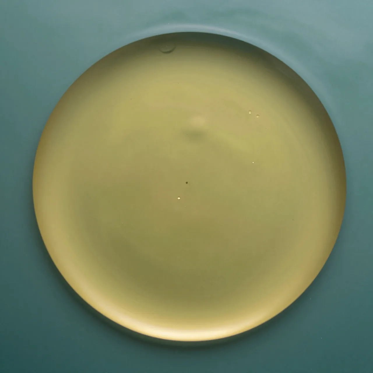 A large smooth golden oil circle floats on a calm teal water surface with tiny trapped air bubbles.