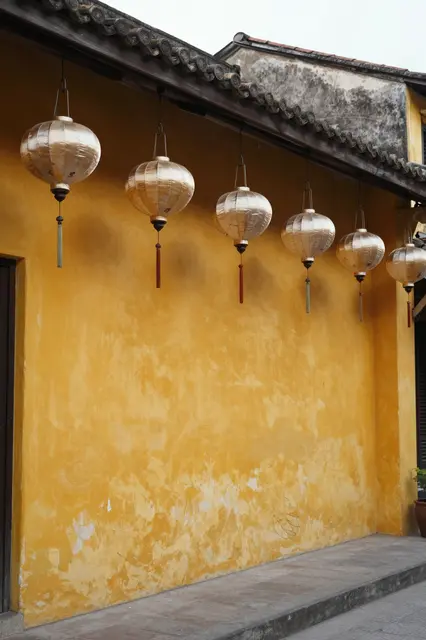 Row of plain pale silk lanterns hangs from the eaves of a weathered yellow plaster building at overcast dusk