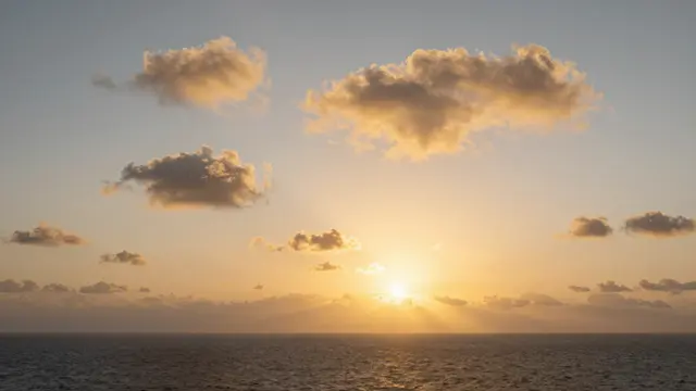 Warm golden sunset light illuminates scattered clouds above a calm dark ocean horizon