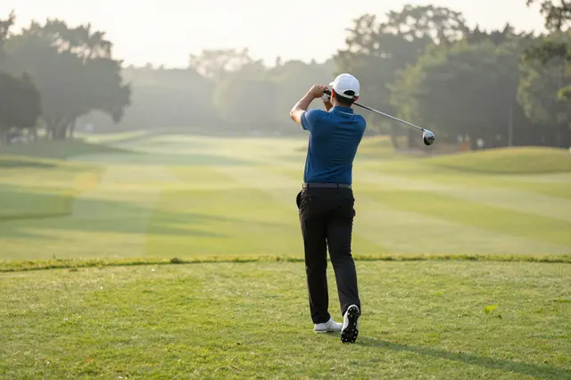 Back view of an adult male golfer mid-swing on a sunlit, hazy golf course at dawn