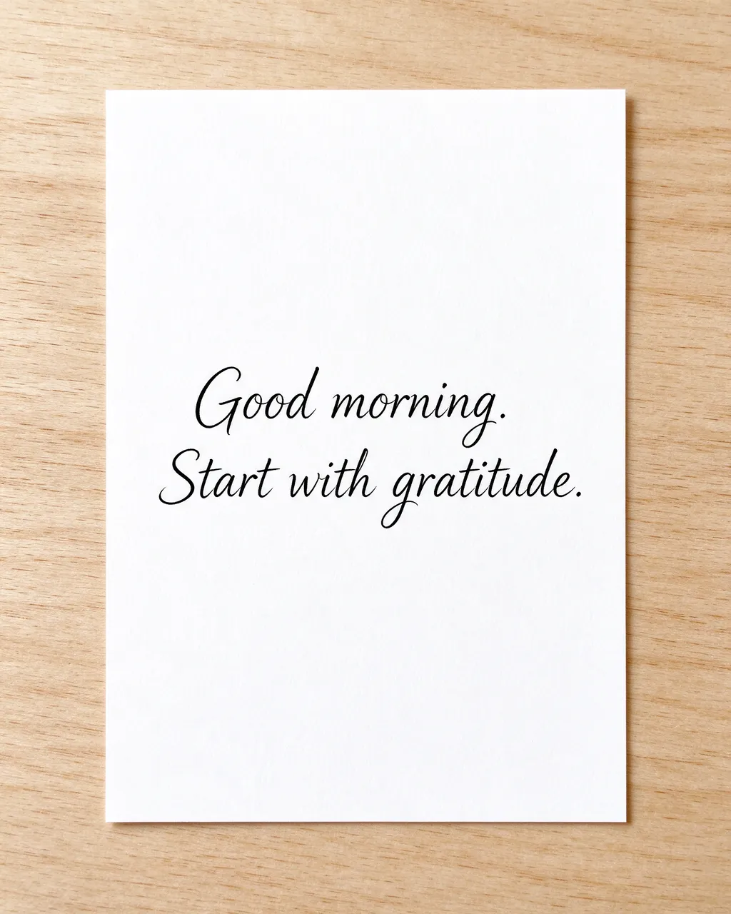 A white index card resting on a light wooden desk displays the text "Good morning. Start with gratitude." in black script